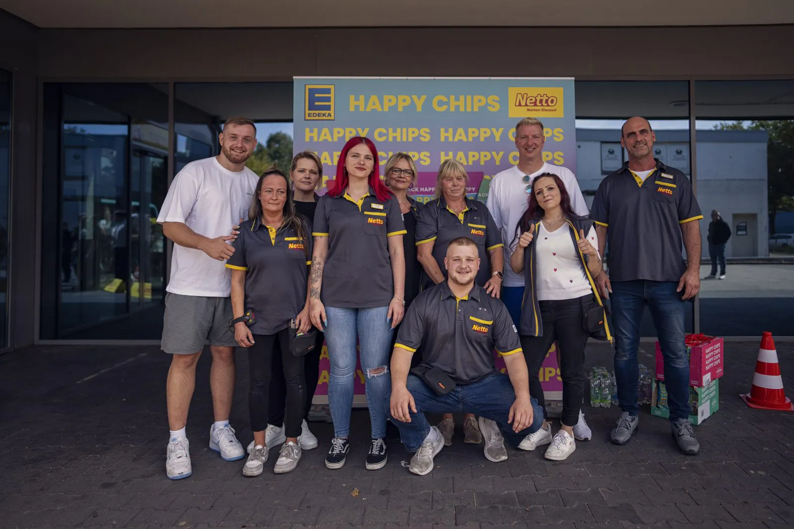 Eine Gruppe von Personen posiert vor einer gelben Happy-Chips-Netto-Wand am Gebäudeeingang.