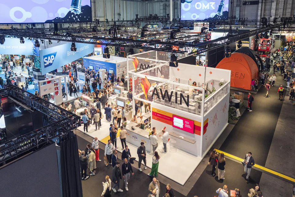 Blick von oben auf den hell beleuchteten Awin-Messestand mit Besuchern in einer großen Messehalle.