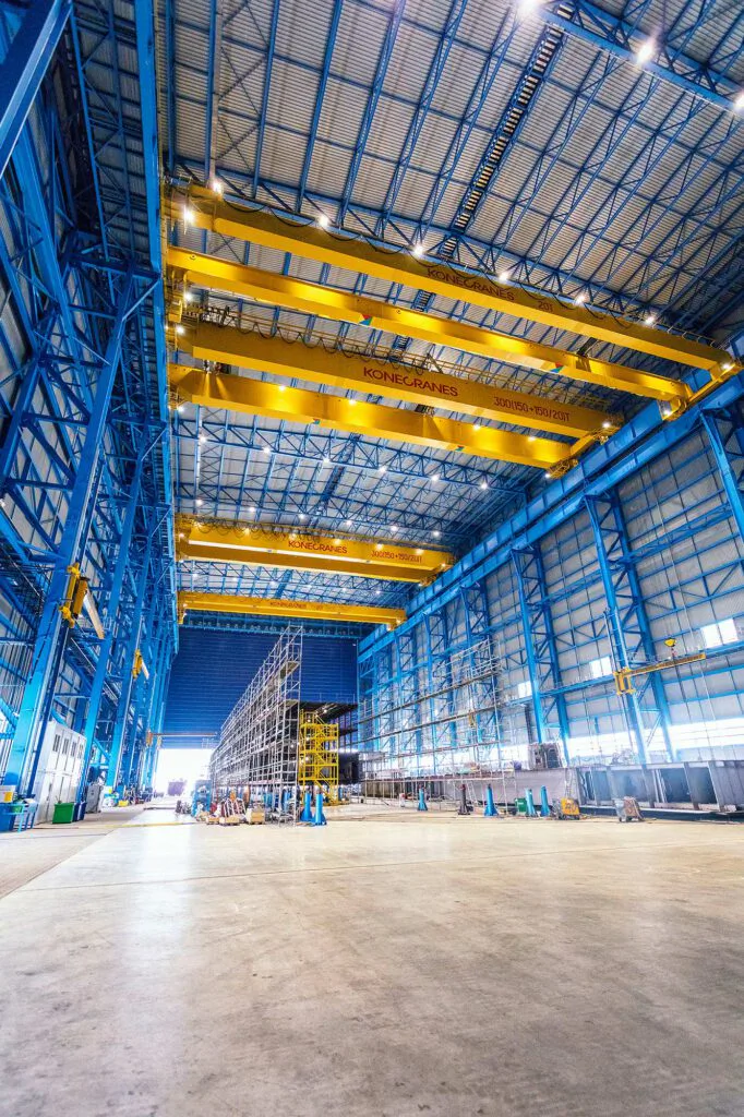Gelbe Konecranes-Brückenkrane unter blauem Stahltragwerk in einer hohen, lichtdurchfluteten Industriehalle.