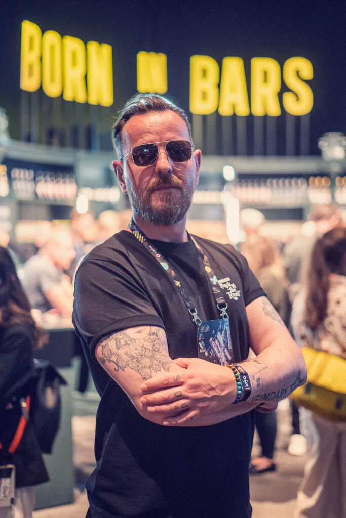 Mann mit Sonnenbrille und tätowierten, verschränkten Armen vor gelbem „Born in Bars“-Schriftzug am Thomas-Henry-Stand.