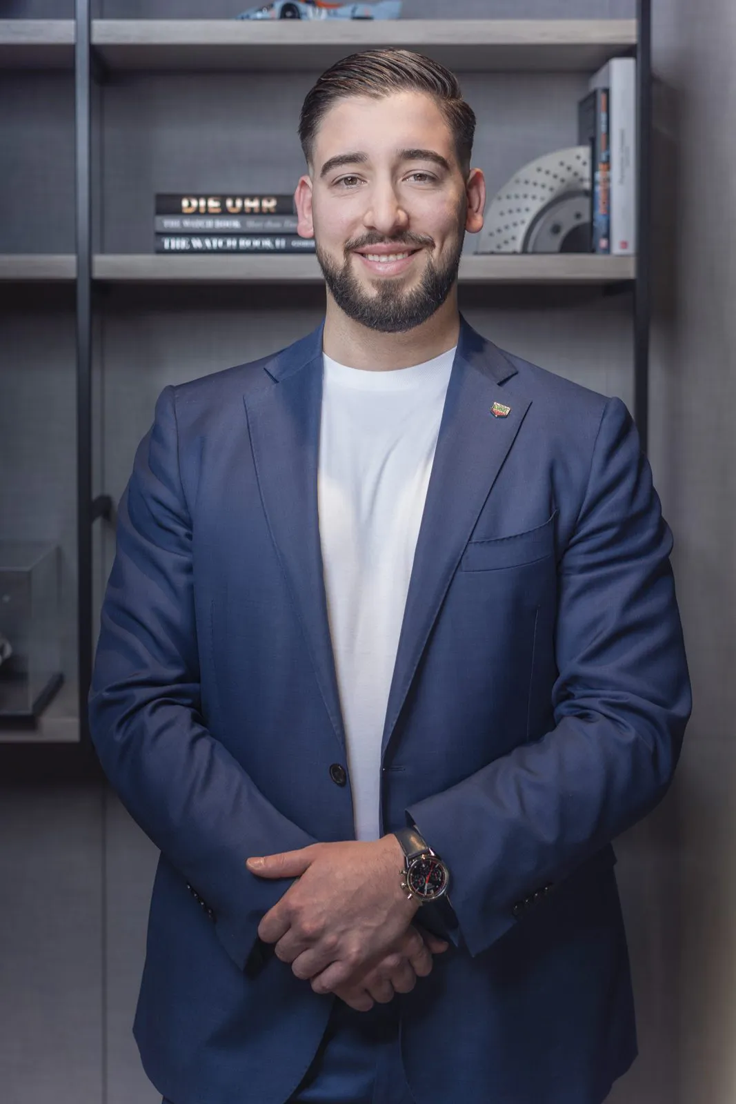 Lächelnder Tag-Heuer-Mitarbeiter im blauen Sakko mit Armbanduhr vor einem Bücherregal, weich beleuchtet.