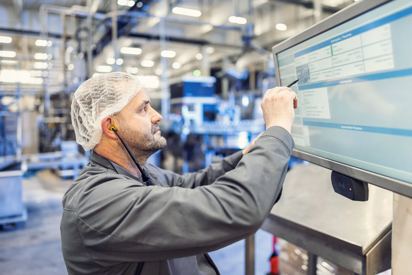 Mitarbeiter mit Haarnetz und Gehörschutz bedient einen Touchscreen in der hell beleuchteten Produktionshalle.