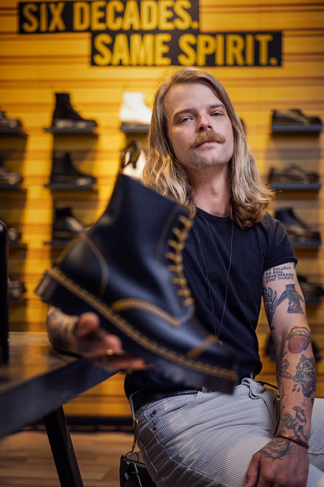 Langhaariger, tätowierter Mann hält einen schwarzen Dr-Martens-Stiefel vor gelber Schuhwand im Laden.