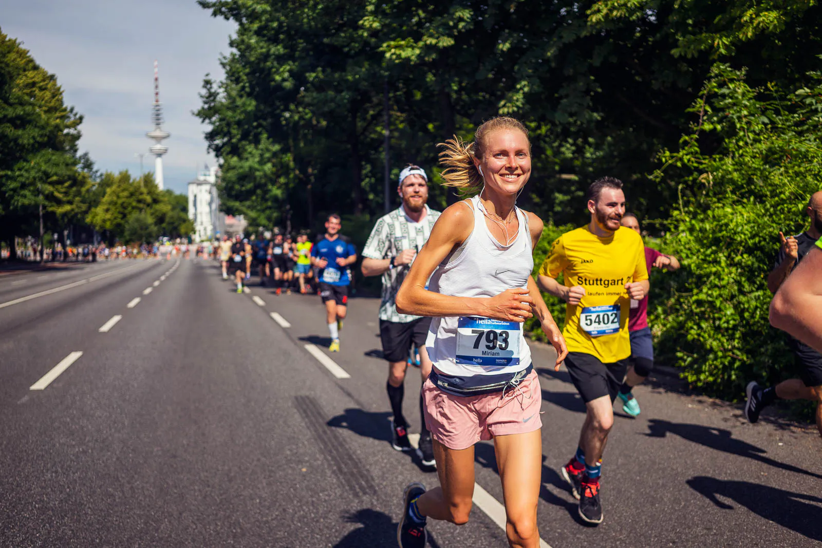 Lächelnde Läuferin mit Startnummer 793 läuft mit weiteren Teilnehmenden auf einer sonnigen, baumgesäumten Straße.