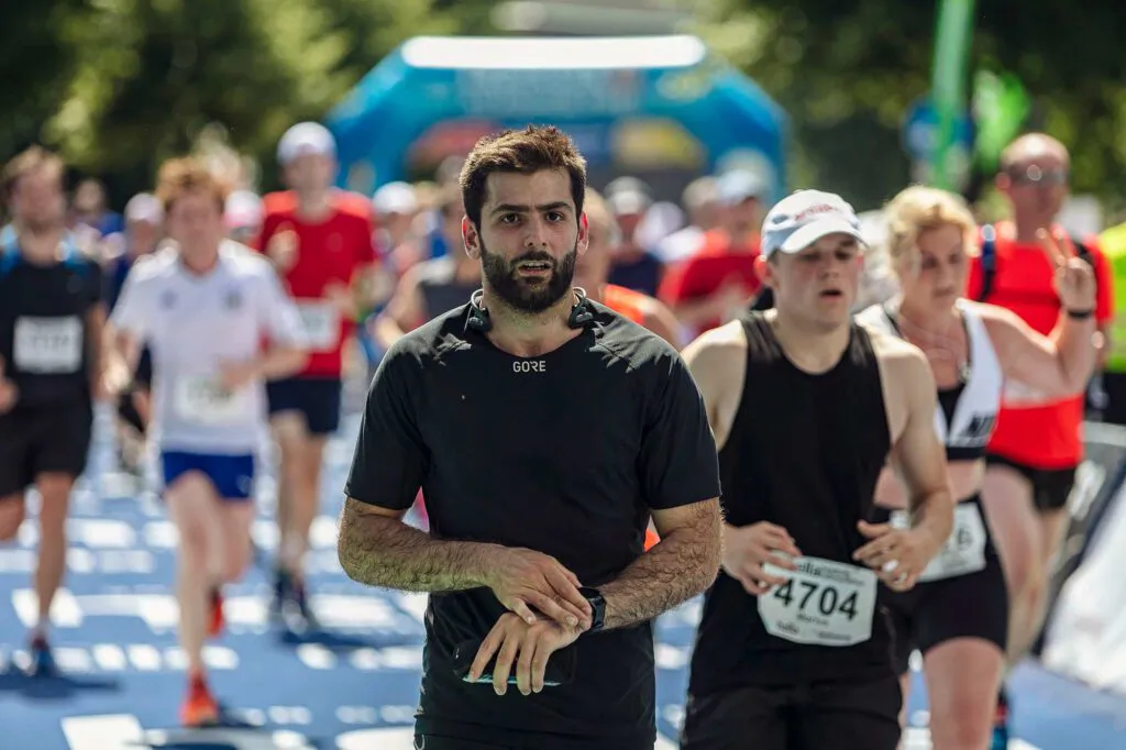 Läufer im schwarzen Shirt greift an seine Sportuhr, umgeben von weiteren Läufern auf sonniger Laufstrecke.