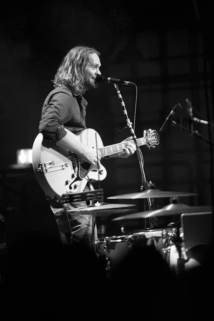 Ingo Pohlmann singt und spielt Gitarre am Mikrofon auf dunkler Bühne, Schlagzeug im Vordergrund, Schwarz-Weiß.