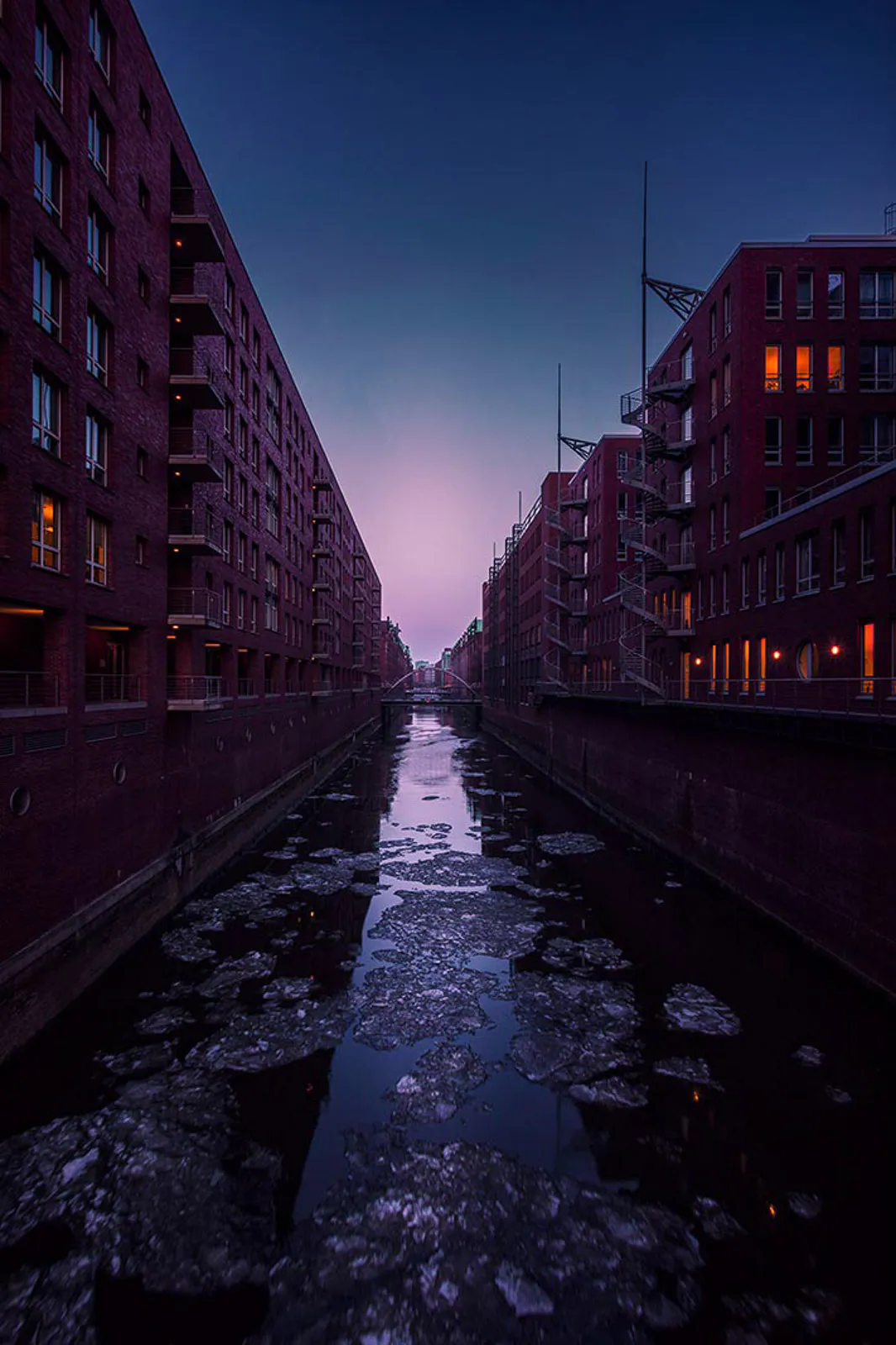 Eisschollen auf einem schmalen Kanal zwischen roten Backsteingebäuden mit beleuchteten Fenstern in der Dämmerung.