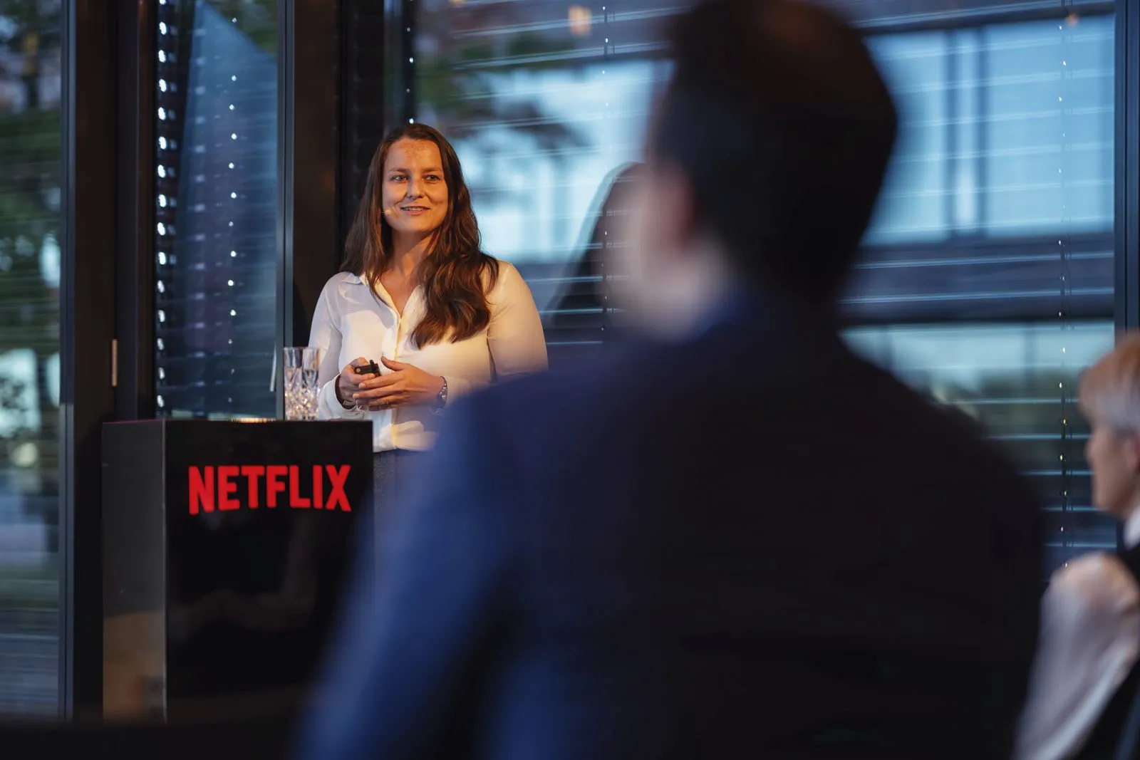 Frau mit Präsentationsklicker hinter einem Pult mit rotem Netflix-Logo, unscharfe Gäste im Vordergrund.