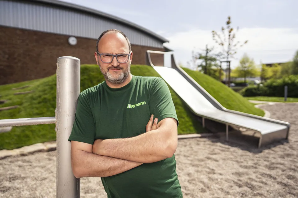 Mann mit Brille und grünem Boymann-Shirt steht mit verschränkten Armen vor einer Rutsche auf einem Spielplatz.