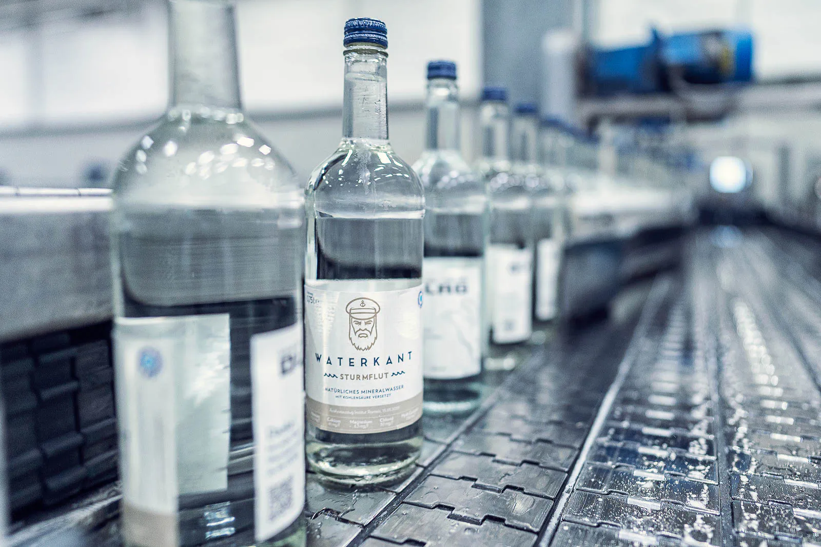 Glasflaschen mit blauen Verschlüssen auf einem nassen Förderband in einer hell beleuchteten Abfüllanlage.