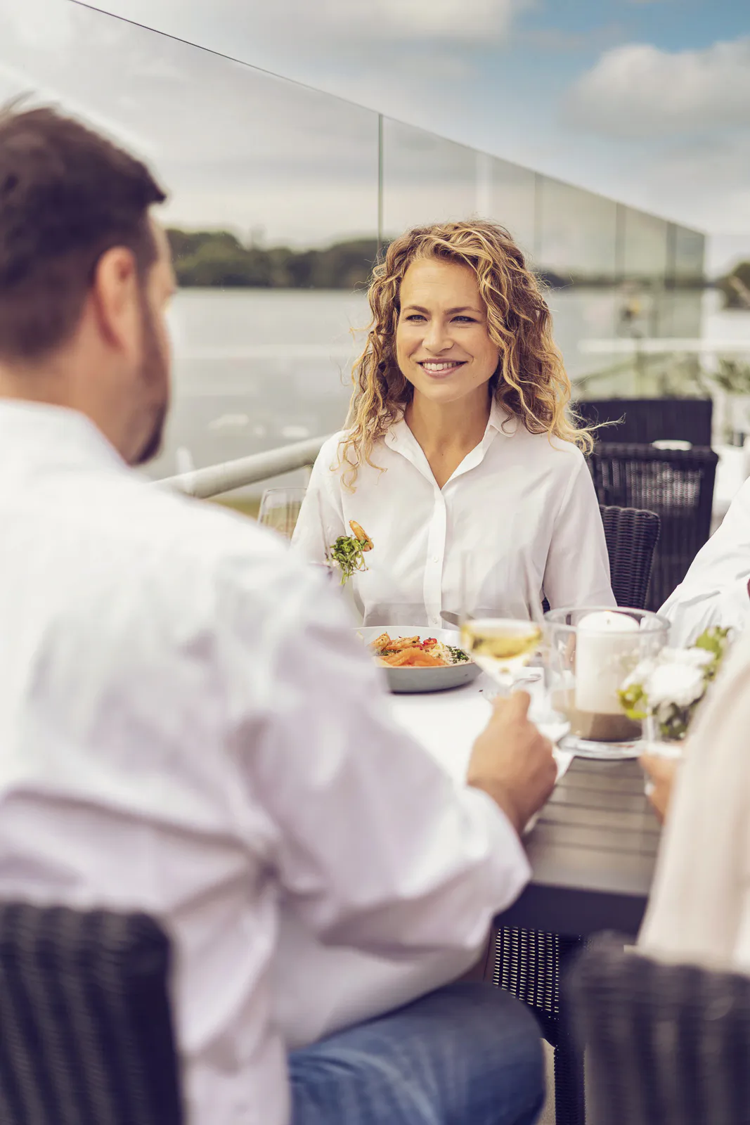 Lächelnde Frau und Mann am gedeckten Tisch mit Essen und Wein auf einer sonnigen Beachclub-Terrasse am Wasser.