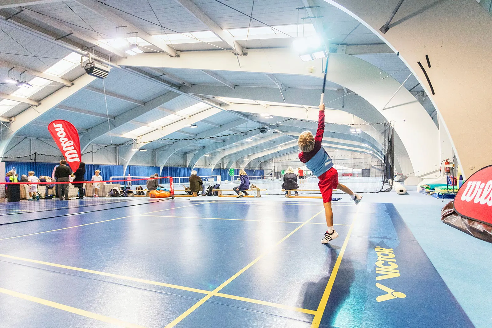 Kind schlägt mit erhobenem Schläger in einer hellen Sporthalle auf, am Spielfeldrand Wilson-Banner.