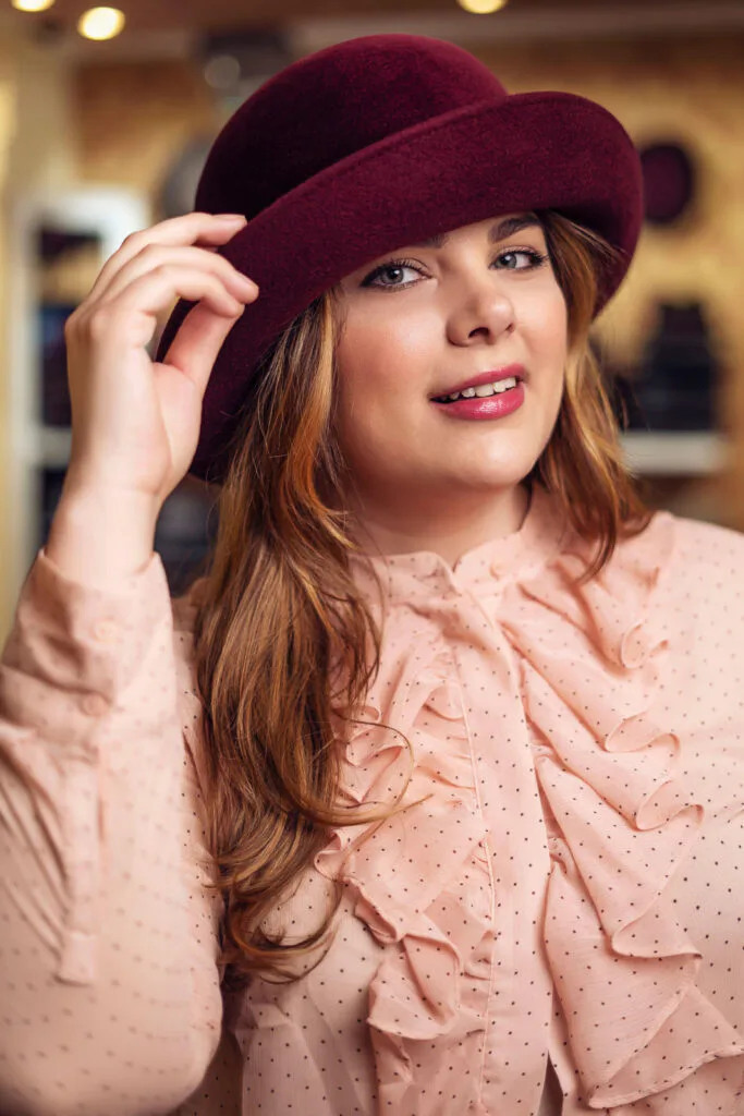 Frau in rosa Rüschenbluse berührt die Krempe ihres bordeauxroten Filzhuts im warm beleuchteten Hutladen.