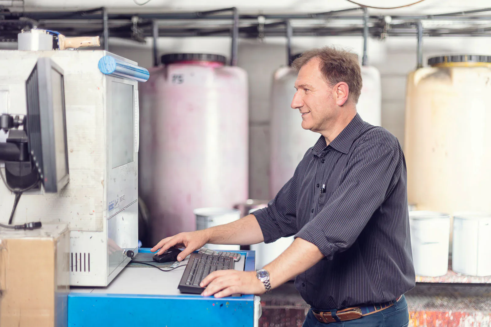 Mitarbeiter bedient einen Computer an einer blauen Arbeitsstation, hinter ihm große Behälter in einer Werkhalle.