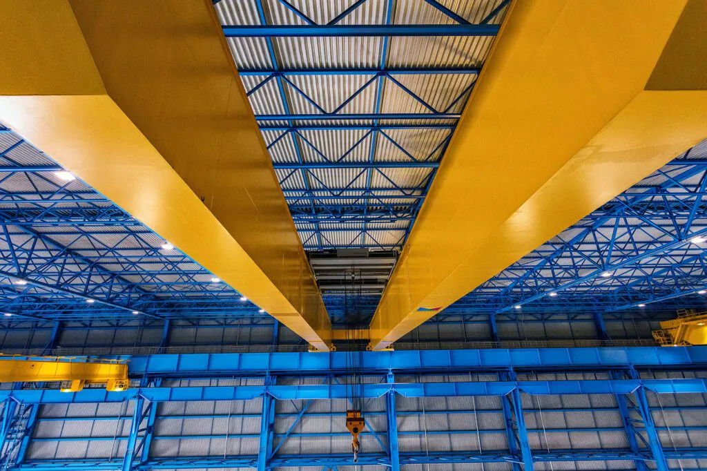 Gelbe Brückenkranträger mit hängendem Kranhaken unter der blau beleuchteten Stahlkonstruktion einer Industriehalle.