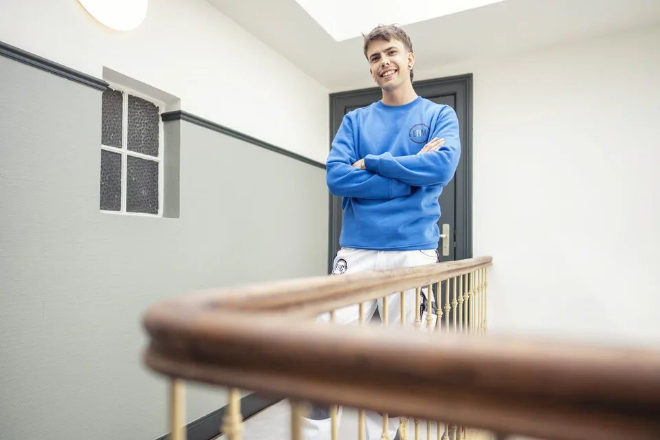 Lächelnder Mann in blauem Pullover mit verschränkten Armen am Holzgeländer eines hellen Treppenhauses.