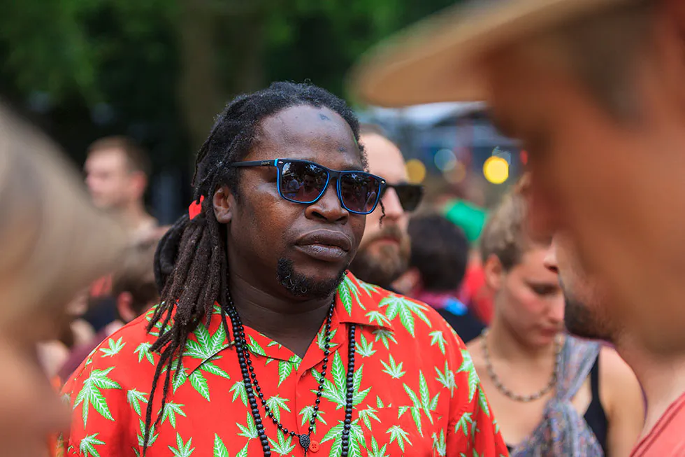 Mann mit Dreadlocks, blauer Sonnenbrille und rotem Hemd mit Blattmuster in einer Menschenmenge im Freien.