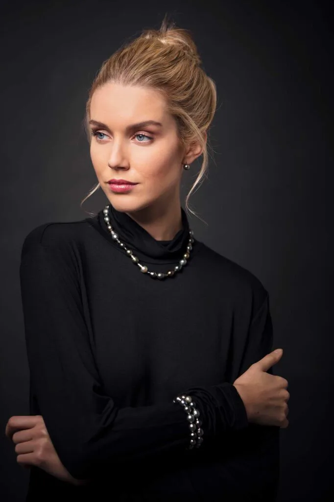 Frau in schwarzem Oberteil mit Perlenkette, Ohrringen und Perlenarmband vor dunklem Studiohintergrund.