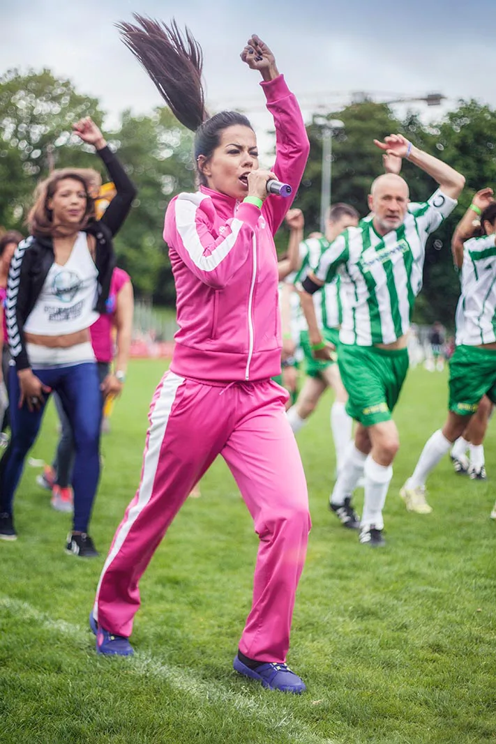 Frau im pinken Trainingsanzug mit Mikrofon und erhobenem Arm auf dem Rasen, dahinter Menschen in Fußballtrikots.
