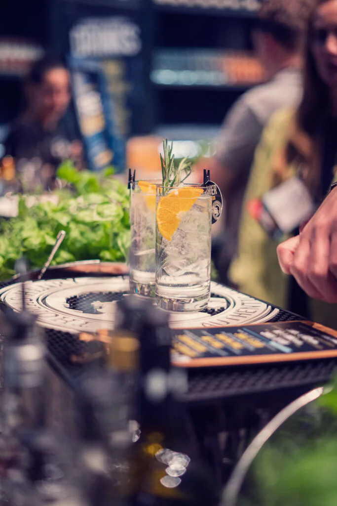 Zwei Drinks mit Eis, Zitruszesten und Rosmarin auf einer Bartheke; Gäste im unscharfen Hintergrund beim OMR Festival.