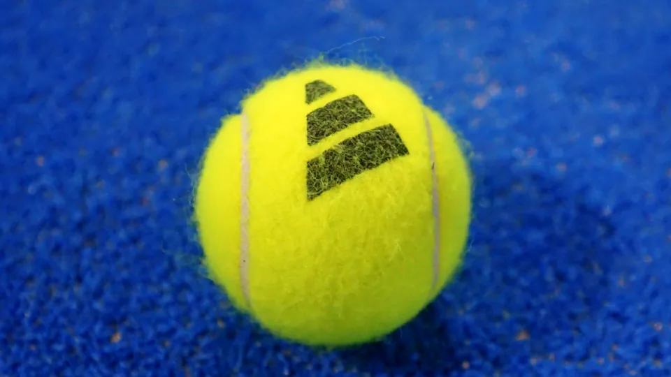 Nahaufnahme eines gelben Padelballs mit schwarzem Logo auf dem blauen Kunstrasen eines Padelplatzes.