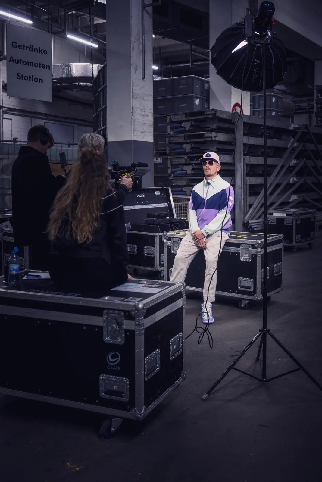 Jan Delay posiert im Trainingsanzug zwischen Equipmentkisten, vor ihm ein Kamerateam und eine Softbox.