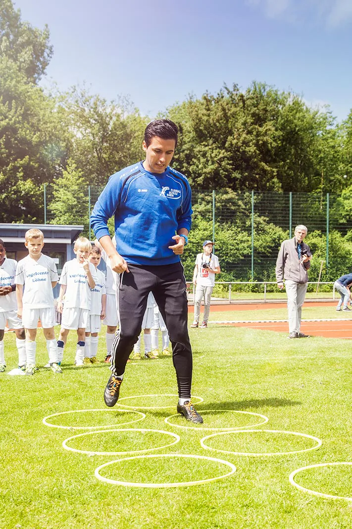 Mann im blauen Sportpullover springt auf sonnigem Rasen durch ausgelegte Reifen, Kinder in Fußballtrikots schauen zu.