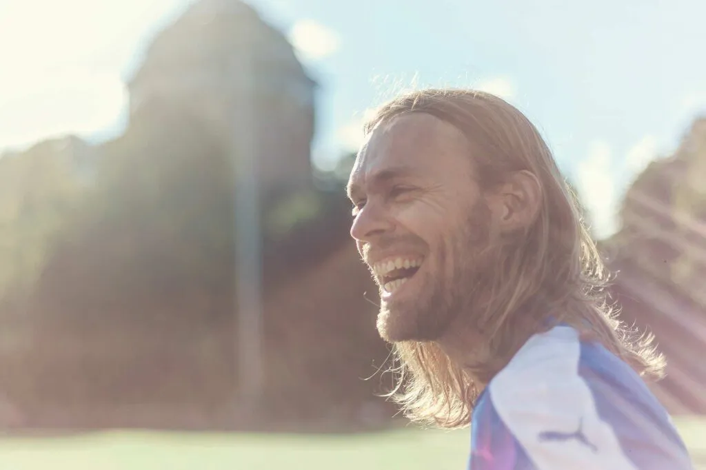 Lachender Mann mit langen Haaren im Fußballtrikot auf einem Sportplatz im warmen Gegenlicht.