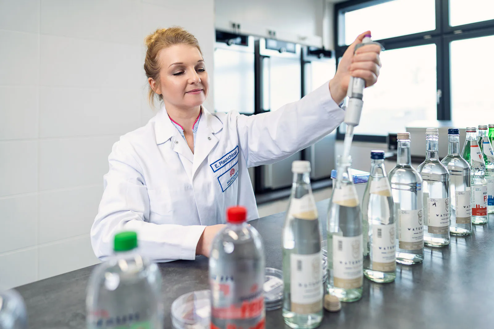 Mitarbeiterin im weißen Kittel pipettiert im hellen Labor eine Wasserprobe; Mineralwasserflaschen stehen auf dem Tisch.