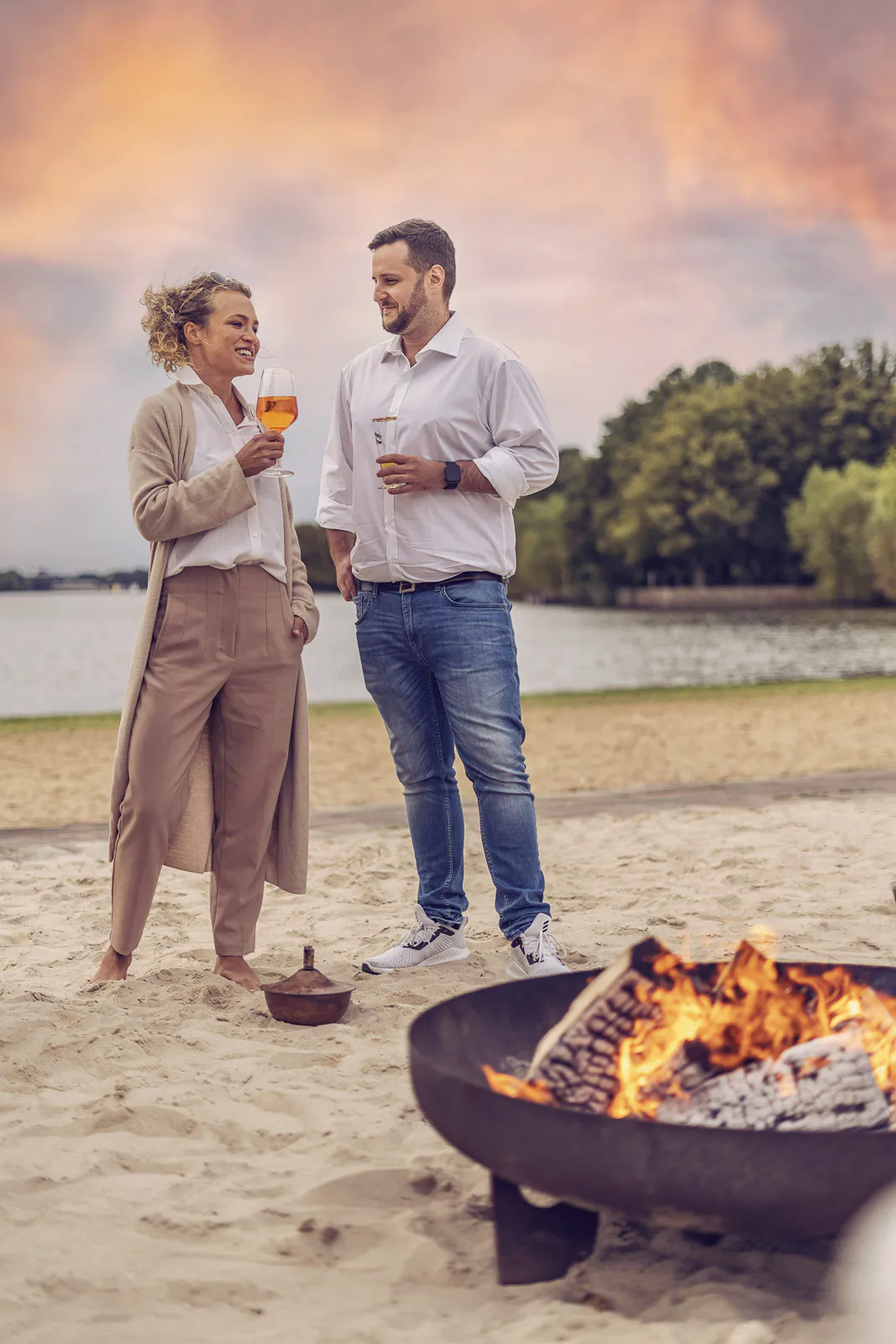 Frau und Mann mit Getränken am Sandstrand neben einer Feuerschale, dahinter ein See im Abendlicht.