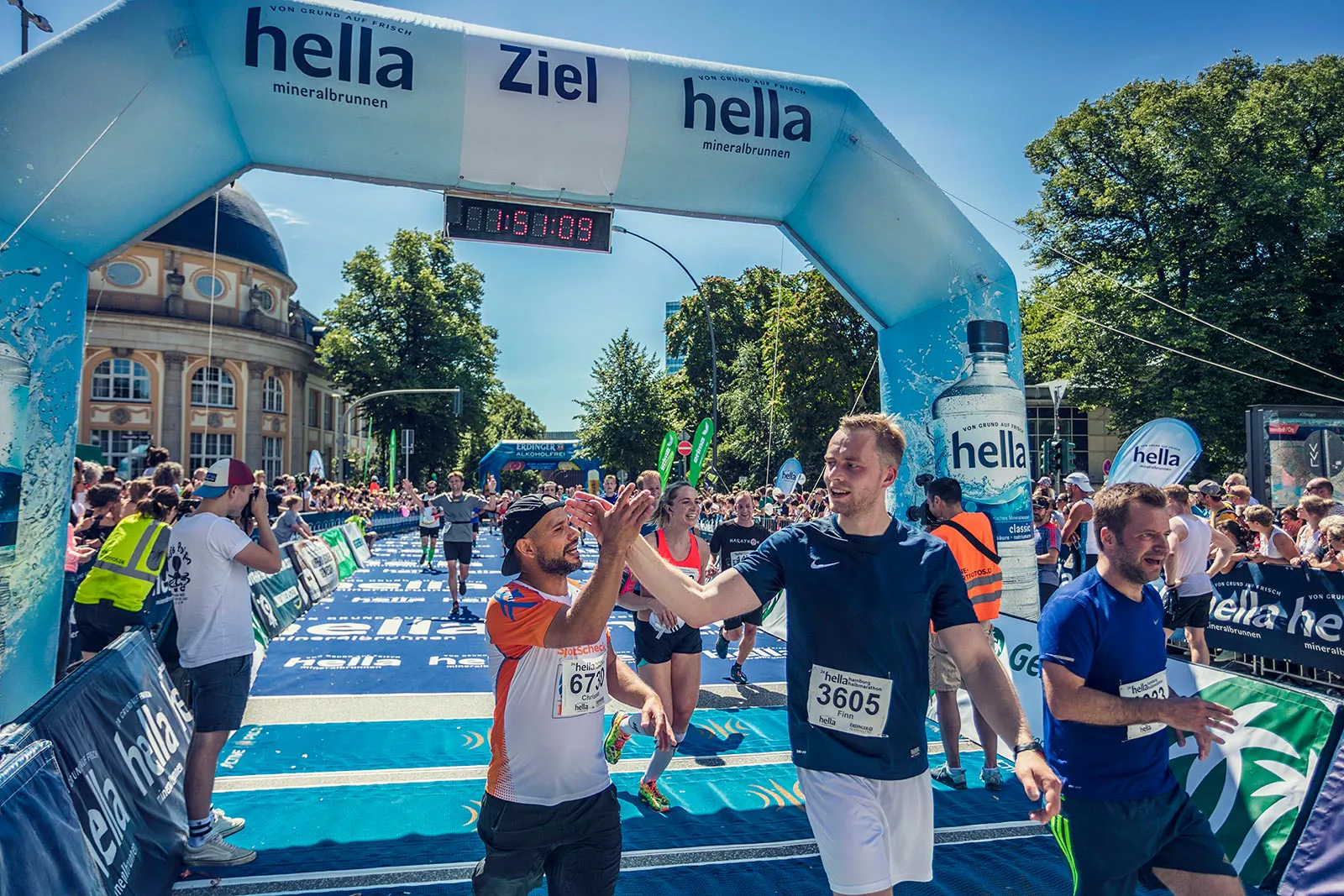 Zwei Läufer klatschen sich im sonnigen Zielbereich des Hella Halbmarathons unter einem blauen Zielbogen ab.