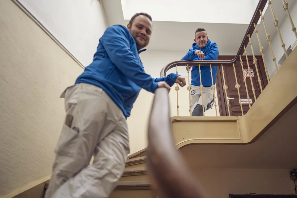 Zwei Handwerker in blauen Jacken und hellen Arbeitshosen lehnen am Geländer eines hellen Altbau-Treppenhauses.