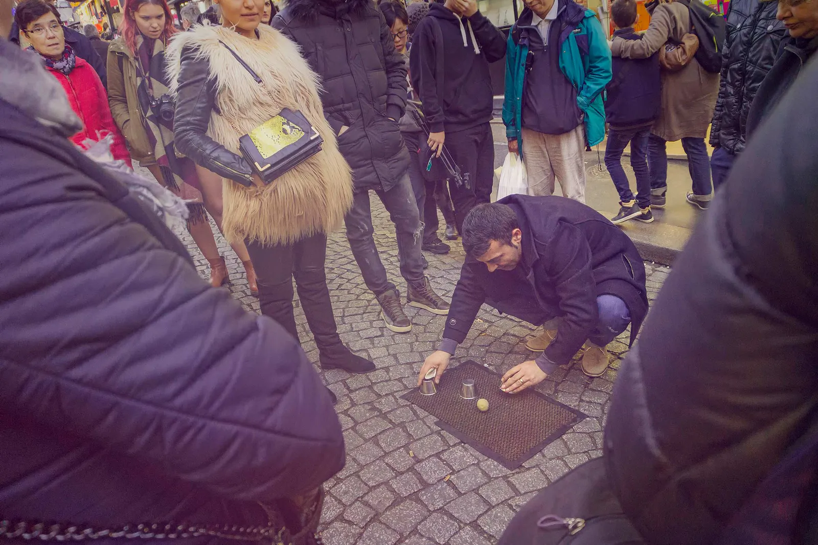 Ein Mann hockt auf Kopfsteinpflaster und spielt vor einer Menschentraube mit Bechern und einem Ball auf einer Matte.