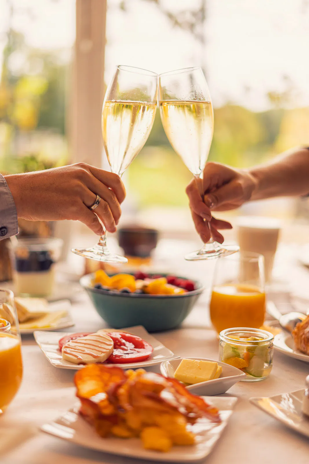 Zwei Personen stoßen im warmen Sonnenlicht mit Sektgläsern über einem Frühstückstisch mit Obst und Orangensaft an.