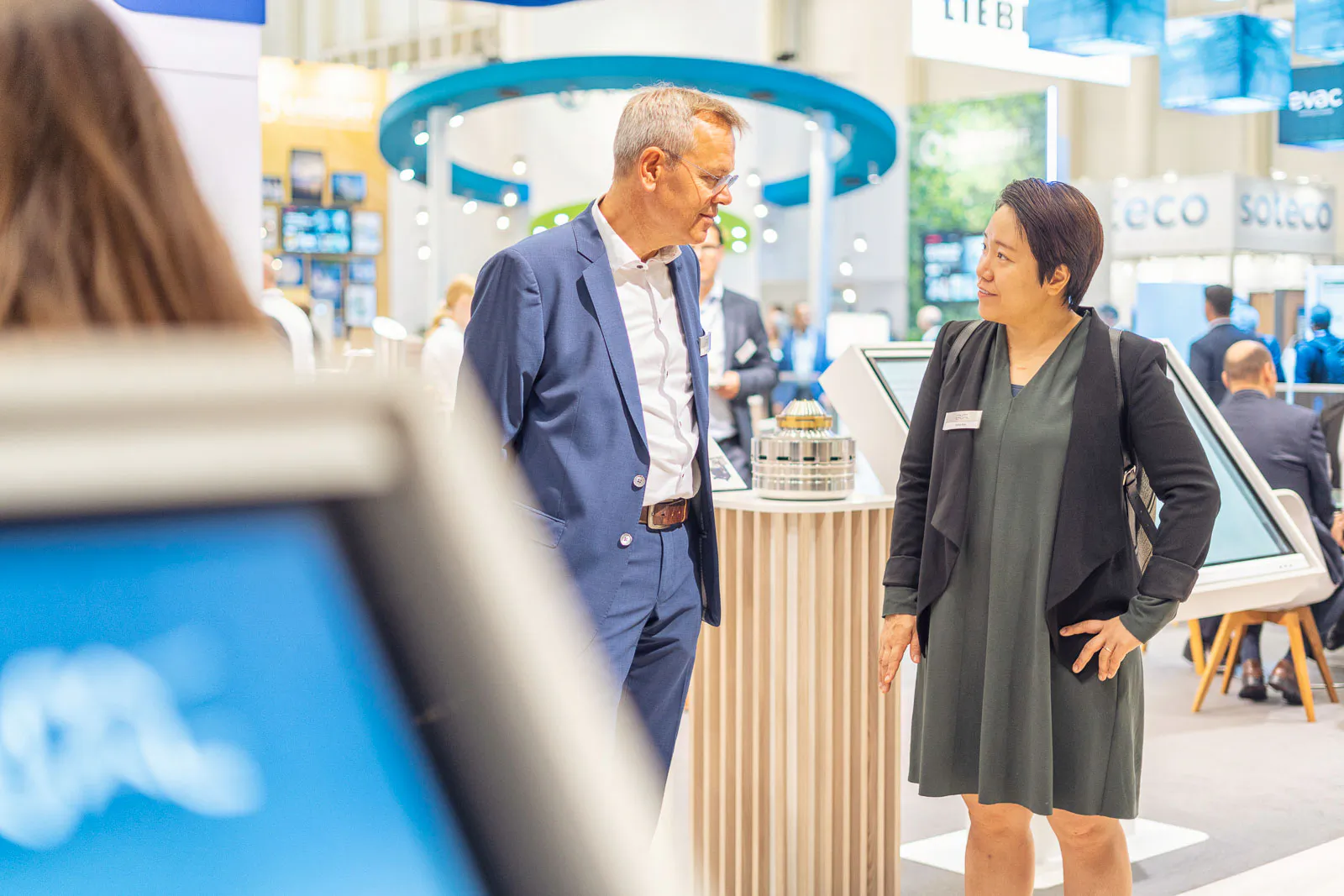 Ein Mann im blauen Anzug und eine Frau im dunklen Kleid sprechen am hell beleuchteten Messestand von Alfa Laval.