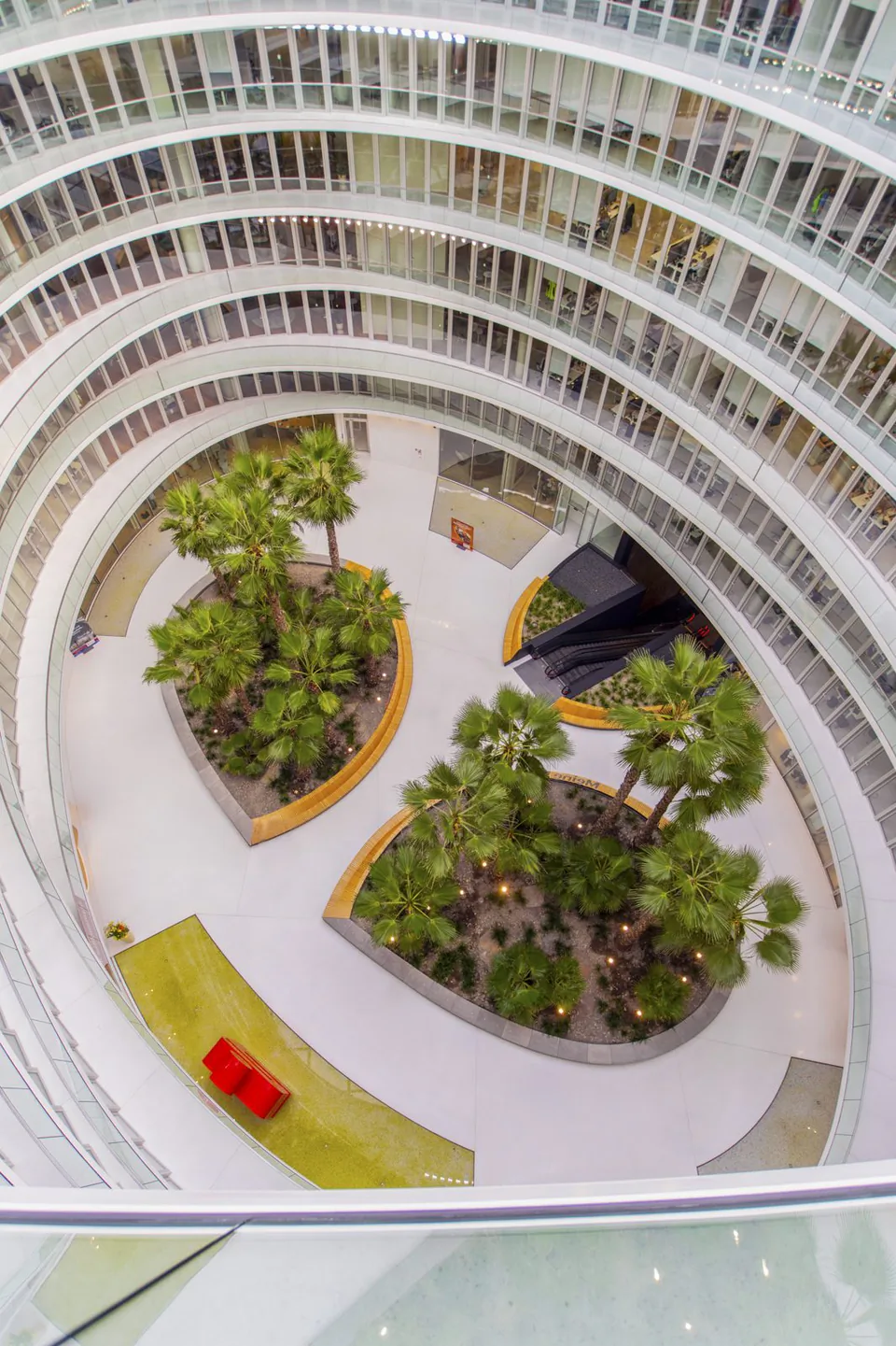 Blick von oben in ein helles, ovales Atrium mit Palmeninseln, roter Sitzbank und umlaufenden Fensterfronten.