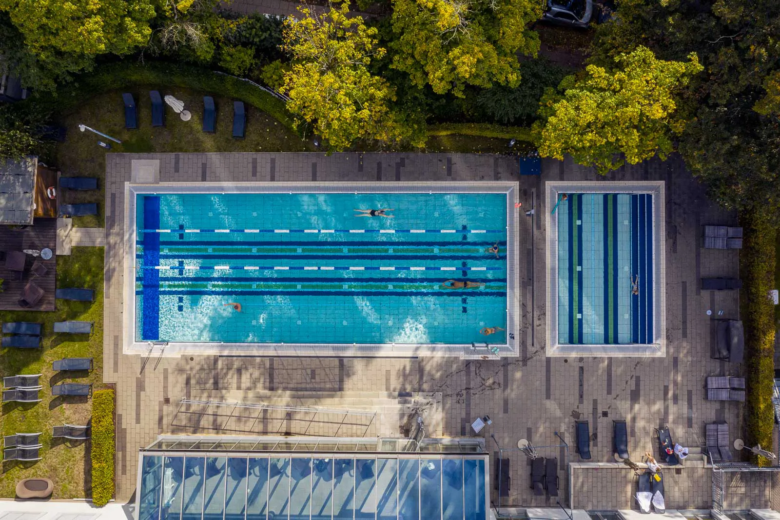 Drohnenaufnahme des Aspria-Außenpools mit Schwimmern auf abgetrennten Bahnen, Liegen und Bäumen im Sonnenlicht.