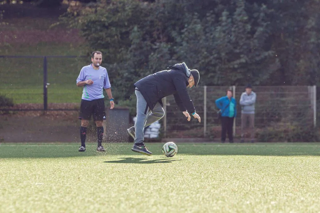 Mann in schwarzer Jacke spielt einen Fußball auf sonnigem Kunstrasen, dahinter stehen ein Spieler und zwei Zuschauer.
