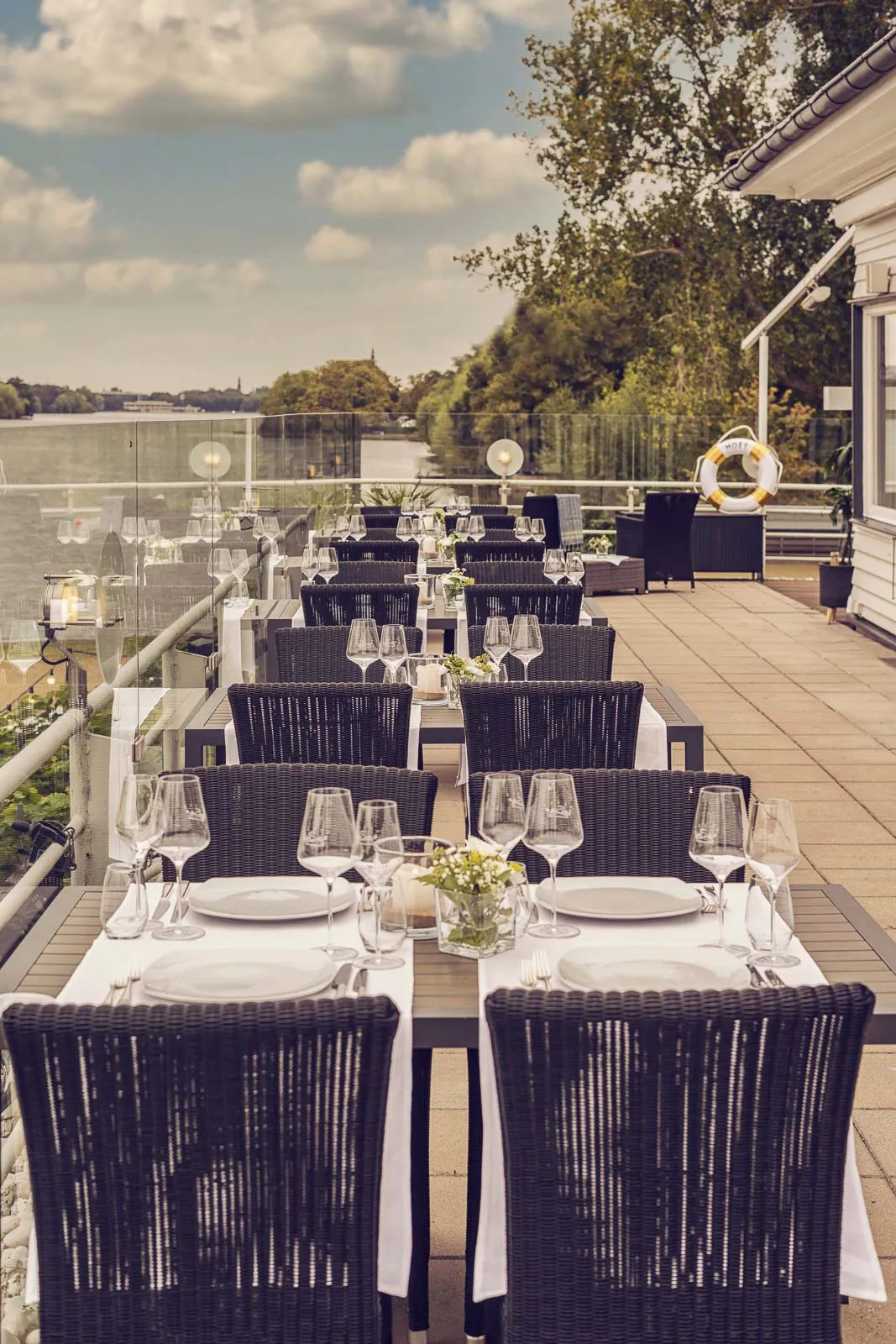 Gedeckte Tische mit Weingläsern und dunklen Korbstühlen auf der sonnigen Terrasse des Insel Beach Club Hannover am See.