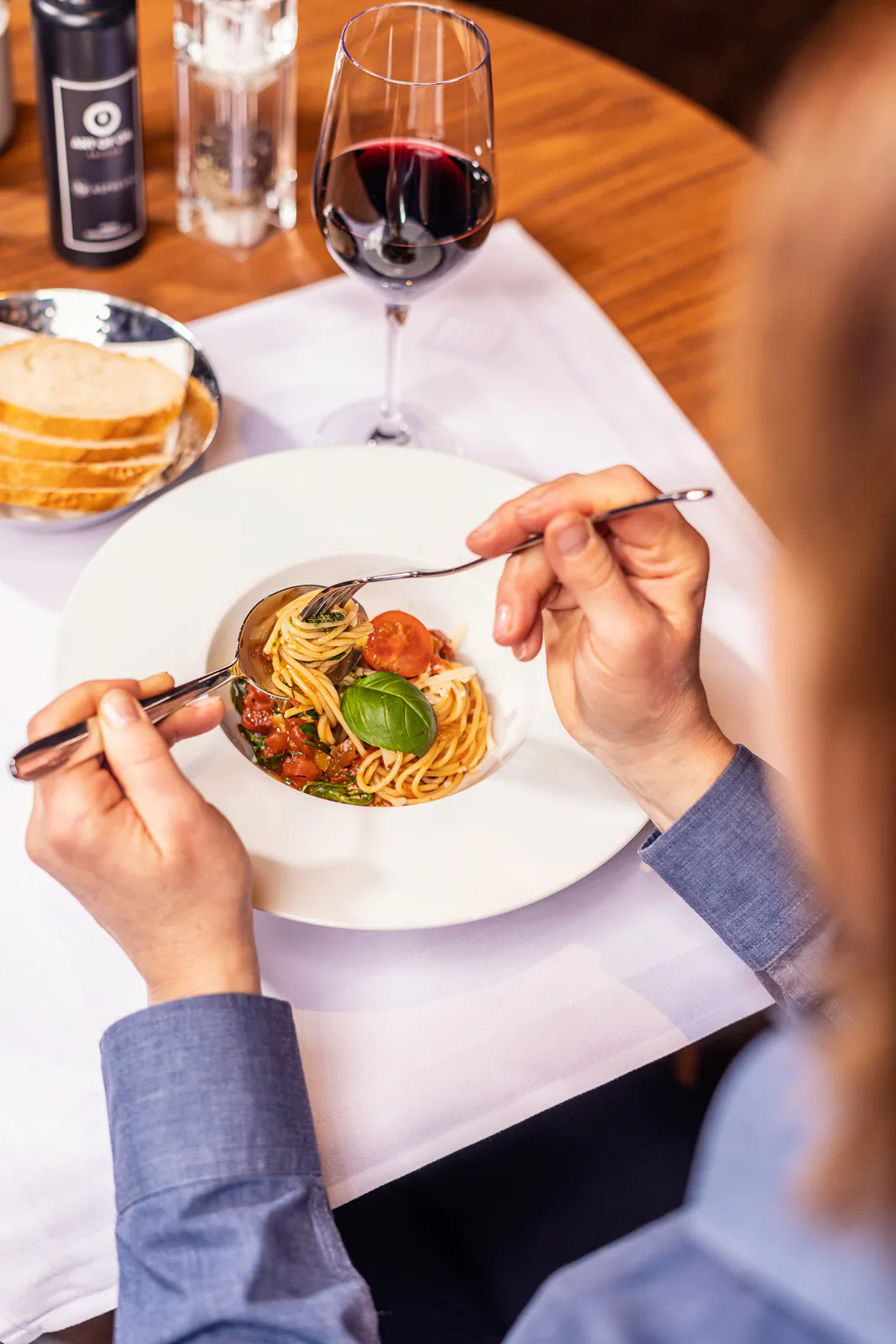 Person isst Spaghetti mit Tomaten und Basilikum im Restaurant, daneben Rotwein und Brot auf dem Holztisch.