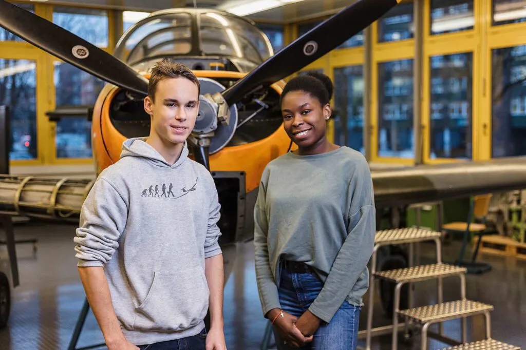 Zwei junge Menschen stehen vor einem orangefarbenen Propellerflugzeug in einer hellen Werkhalle.