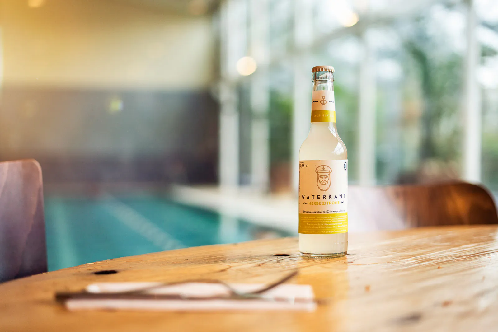 Eine Flasche Waterkant steht auf einem Holztisch im hellen Restaurant, davor liegen Besteck und Serviette.
