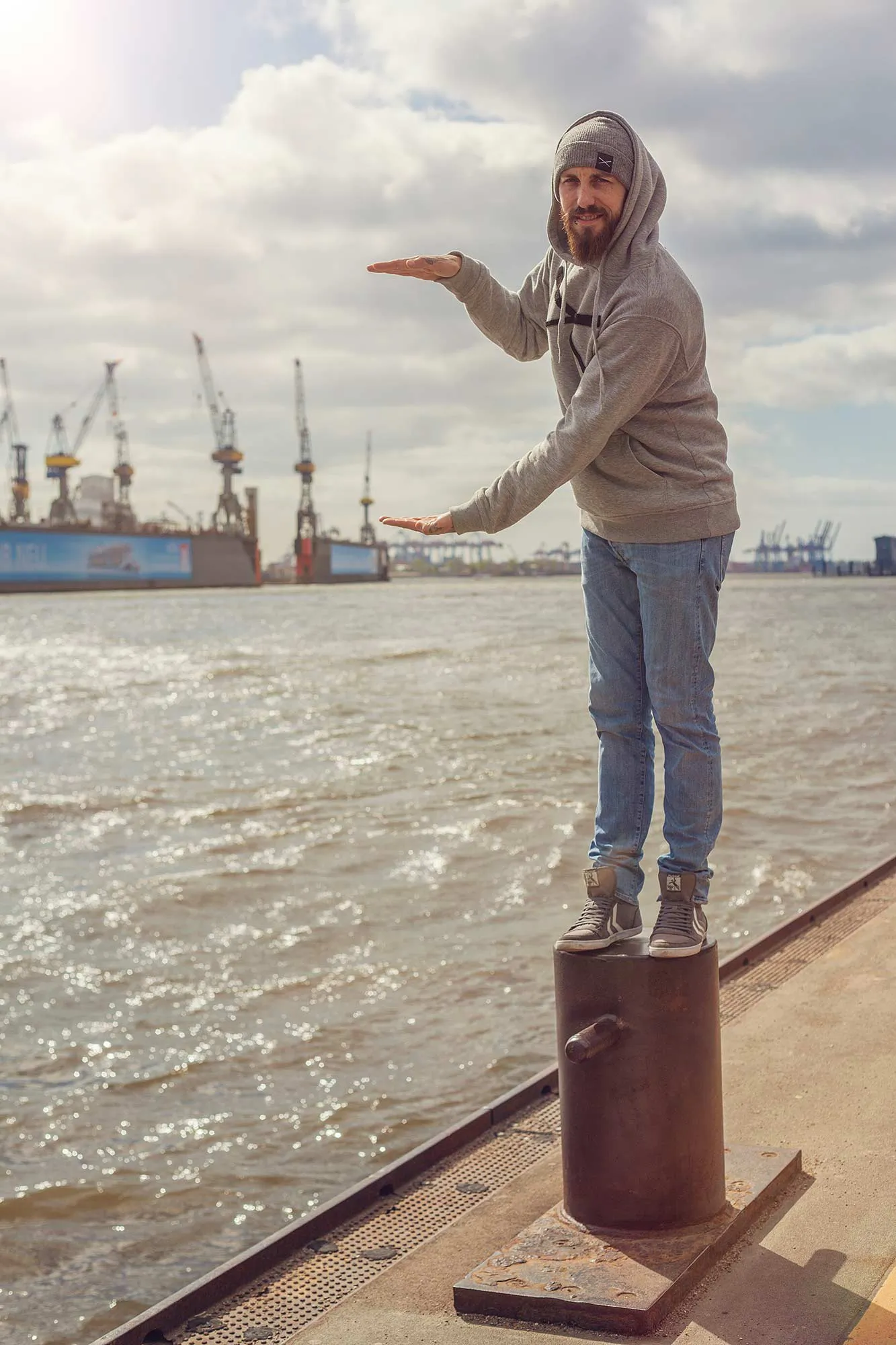 Mann im grauen Hoodie steht auf einem Poller am sonnigen Hafenkai und hält die Hände vor Kräne im Hintergrund.
