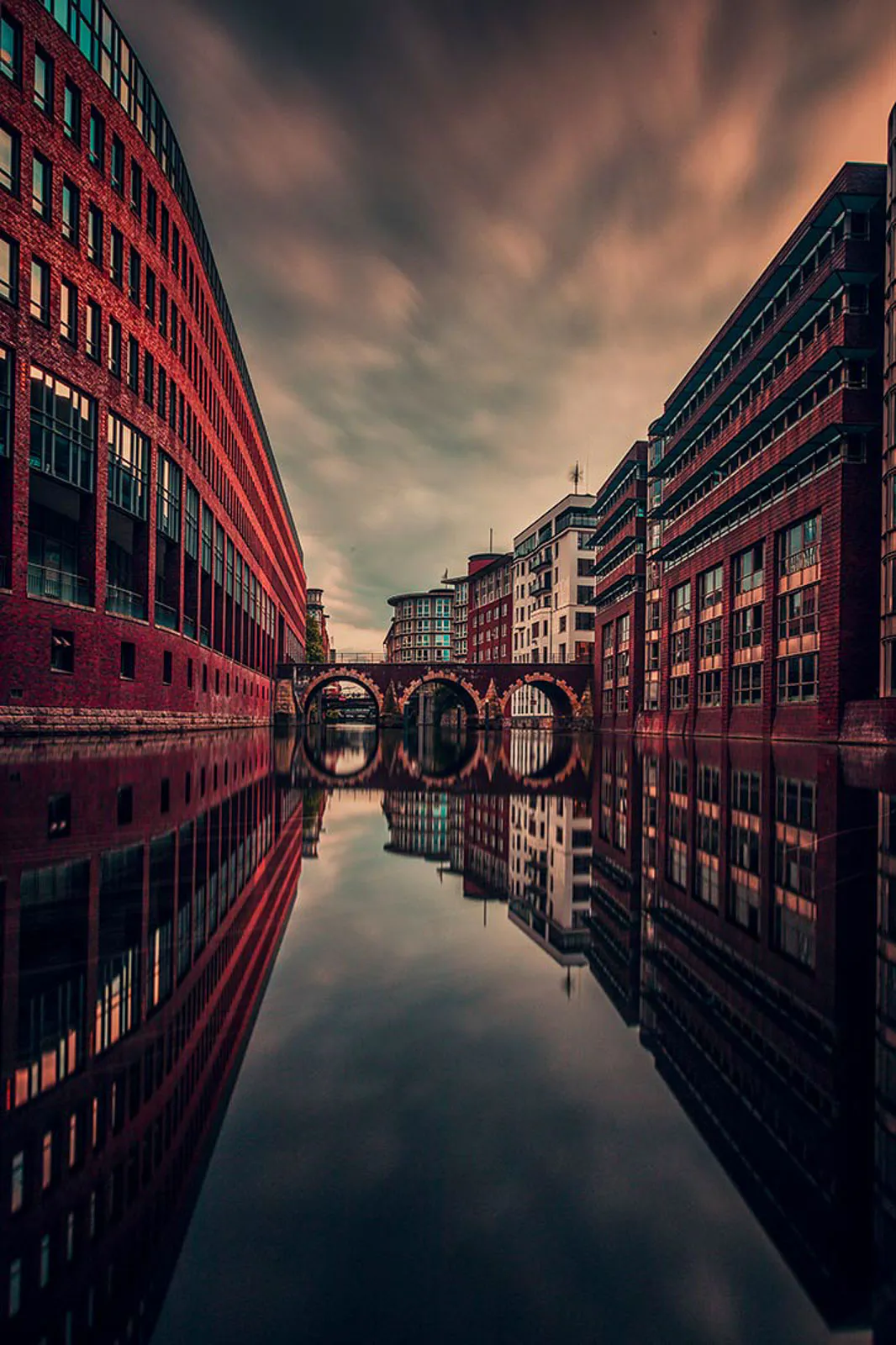 Backsteingebäude und eine Bogenbrücke spiegeln sich im stillen Kanal unter rötlich leuchtendem Wolkenhimmel.
