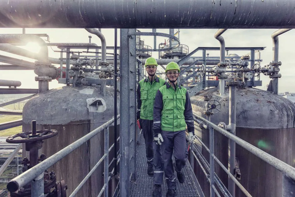 Zwei Arbeiter mit grünen Helmen gehen im Gegenlicht über einen Steg zwischen Rohren und Behältern der Raffinerie Heide.