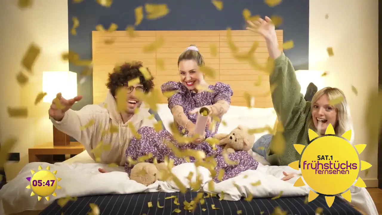Drei lachende Personen auf einem Bett mit Teddybären und goldenem Konfetti im hellen Sat. 1 Frühstücksfernsehen-Set.