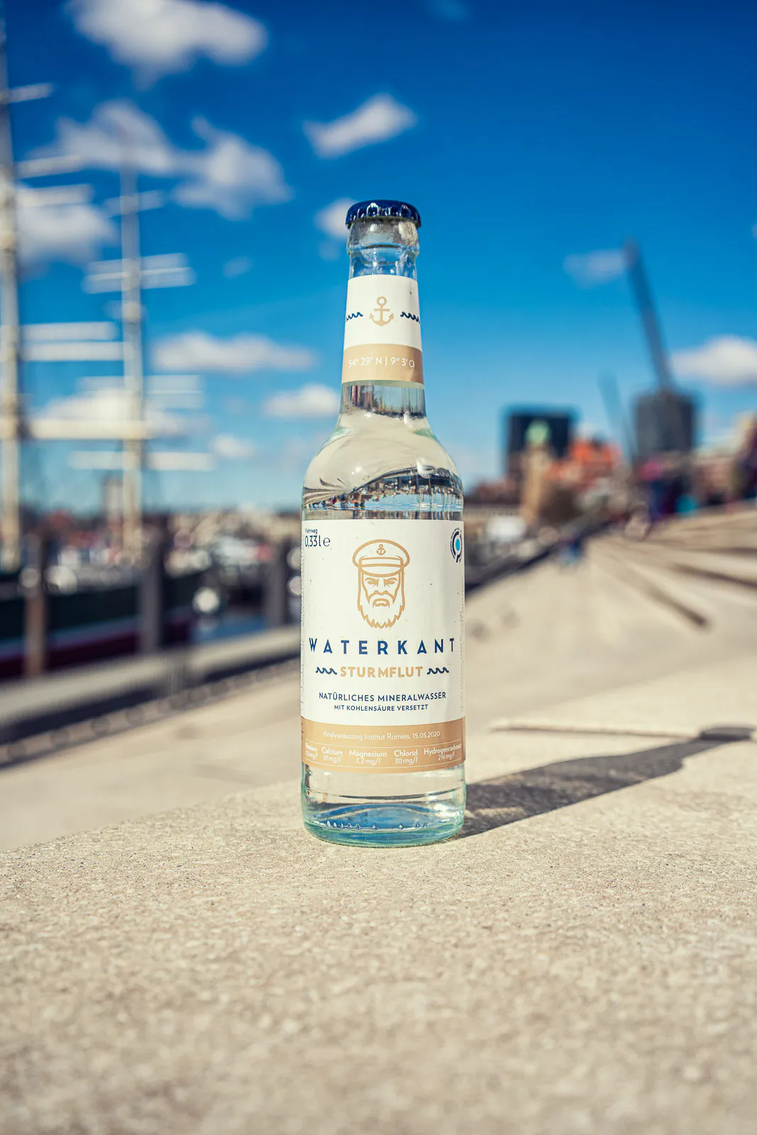 Waterkant-Mineralwasserflasche auf einer sonnigen Steinmauer am Hafen, mit Segelmasten und blauem Himmel im Hintergrund.