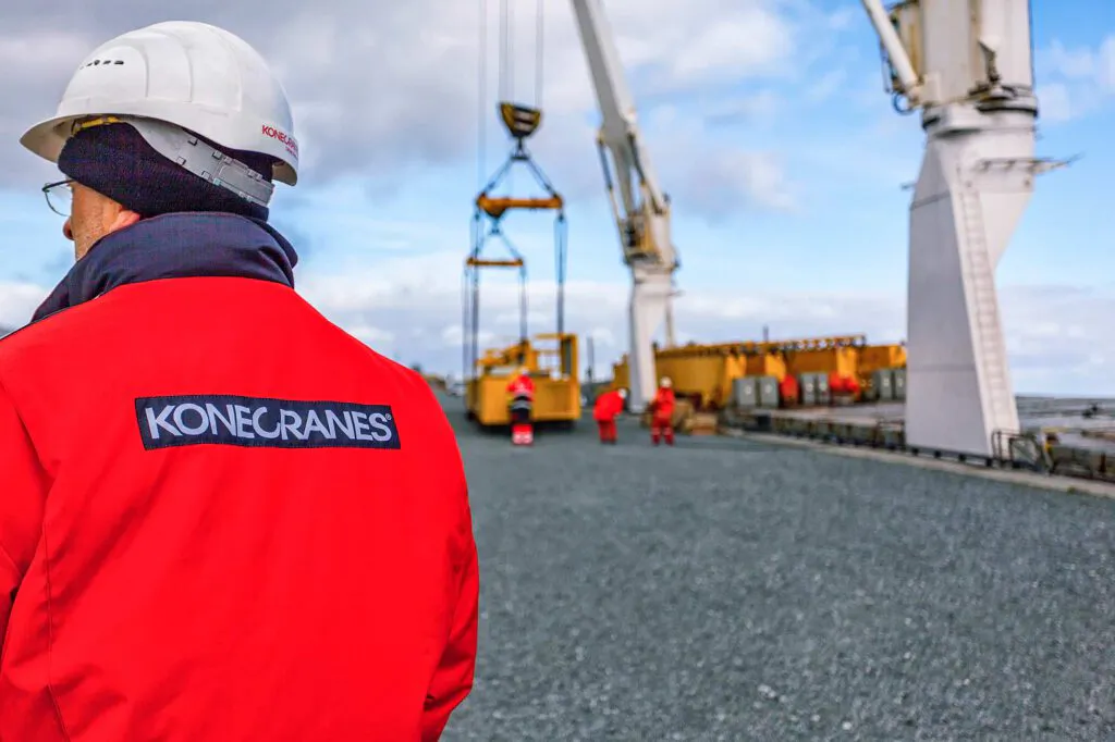 Konecranes-Mitarbeiter mit weißem Helm und roter Jacke vor Hafenkränen und schwebender Last in Cuxhaven.