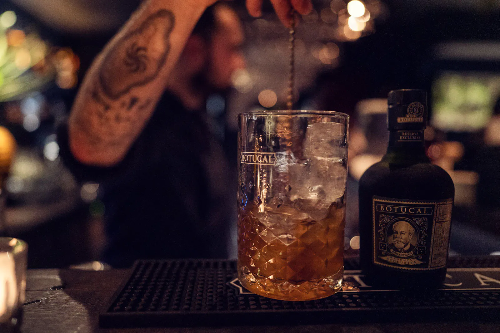 Tätowierte Person rührt einen Drink mit Eis im Mixglas neben einer BOTUCAL-Rumflasche an einer warm beleuchteten Bar.