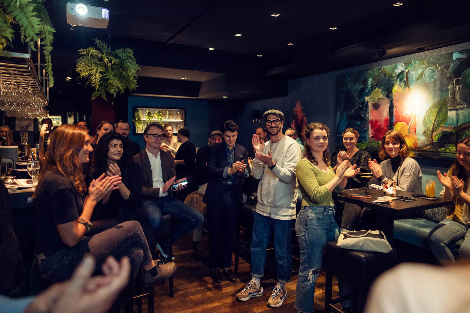 Gäste applaudieren in einer stimmungsvoll beleuchteten Bar mit Hängepflanzen und Getränken auf Stehtischen.